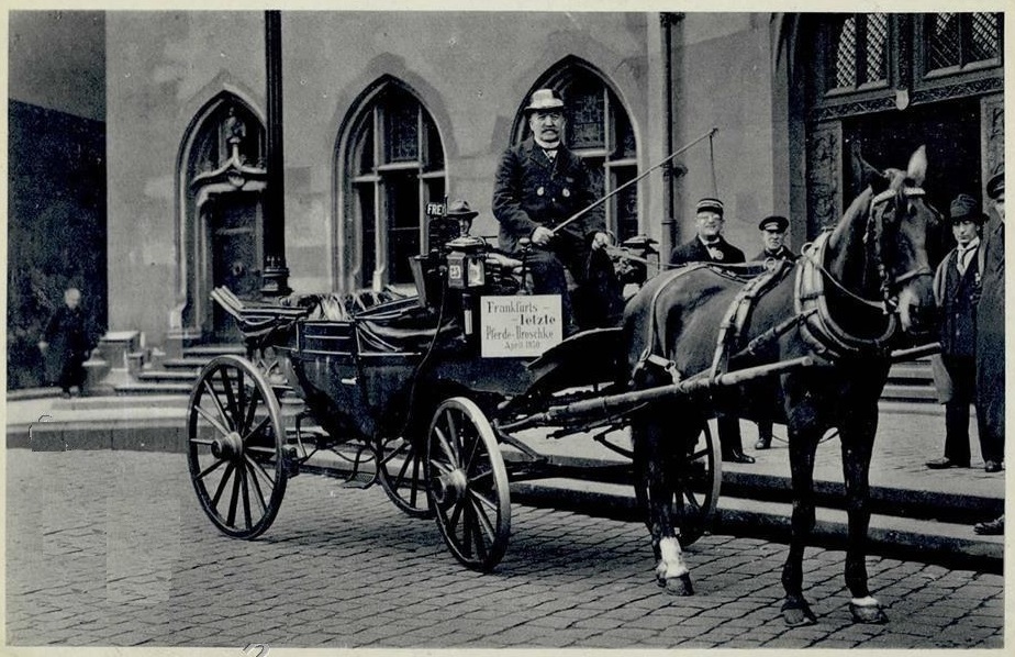 Vor einem gotischen Gebäude steht eine Pferdedroschke mit Kutscher auf dem Bock. Am Wagen ein Schild: „Frankfurts letzte Pferde-Droschke, April 1930“. Umstehende Männer, Kopfsteinpflaster.
