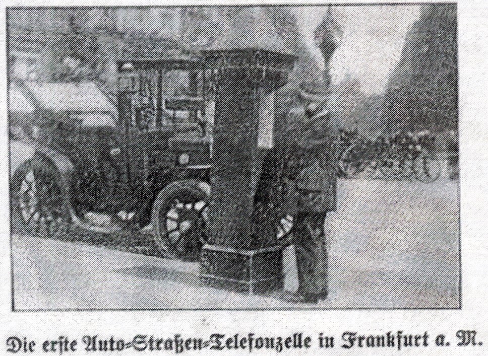 Straßenaufnahme: eine gemauerte Telefonsäule am Straßenrand, davor ein Mann in Uniform am Hörer, dahinter eine offene Motordroschke.