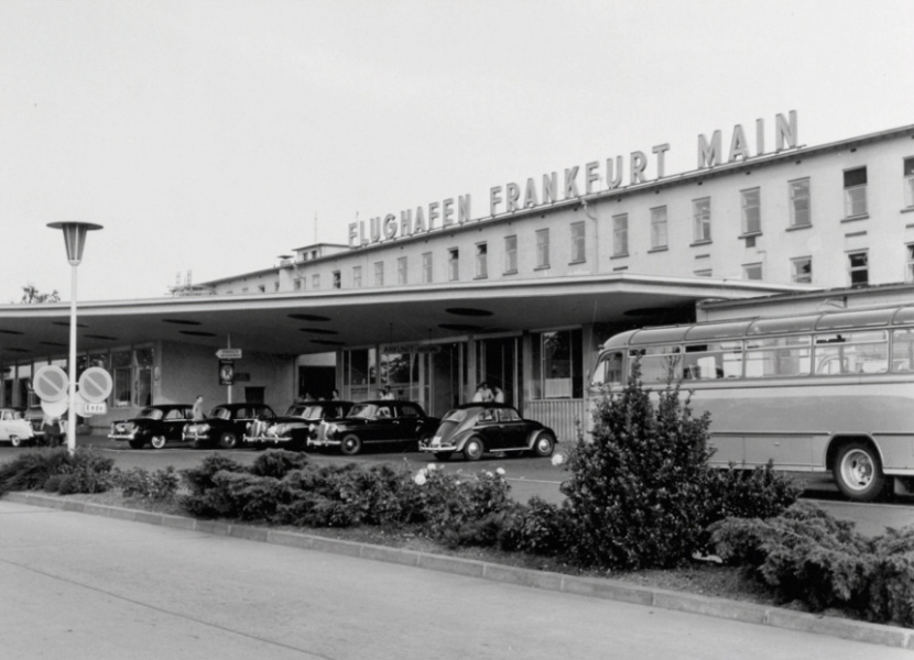 Das alte Empfangsgebäude des Flughafens mit dem Schriftzug „Flughafen Frankfurt Main“. Unter dem Vordach eine Reihe wartender Taxis, rechts ein Reisebus, davor eine Grünanlage.
