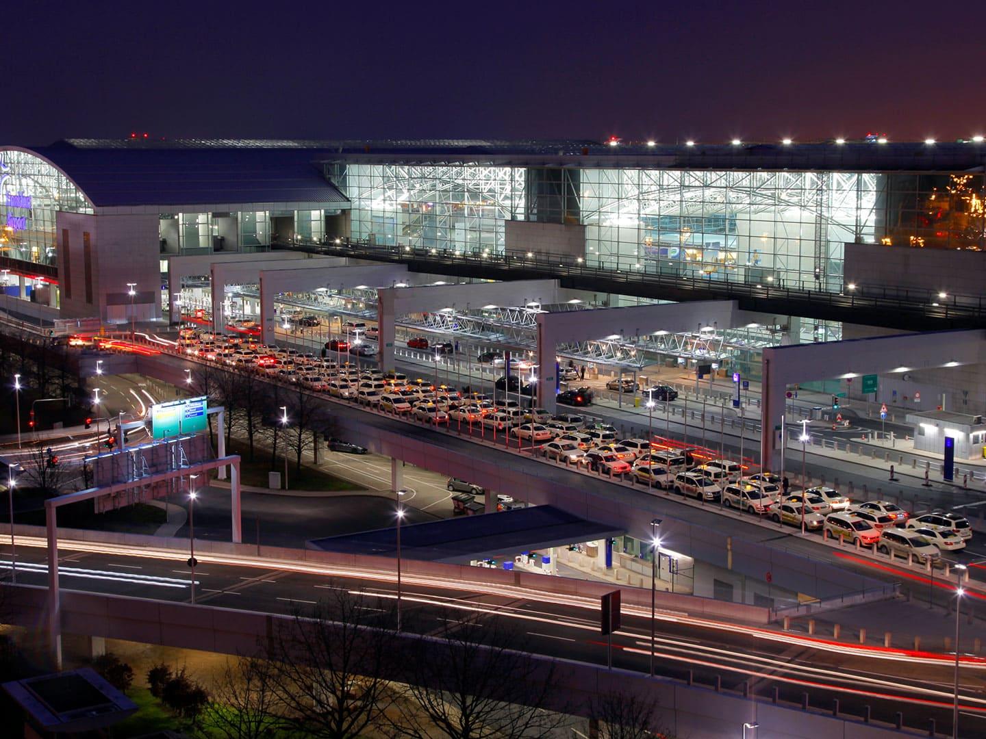 Nachtaufnahme des Flughafens Frankfurt: die beleuchtete Glasfassade von Terminal 1, davor auf mehreren Spuren eine lange Reihe wartender Taxis, darunter Fahrbahnen mit Lichtspuren.