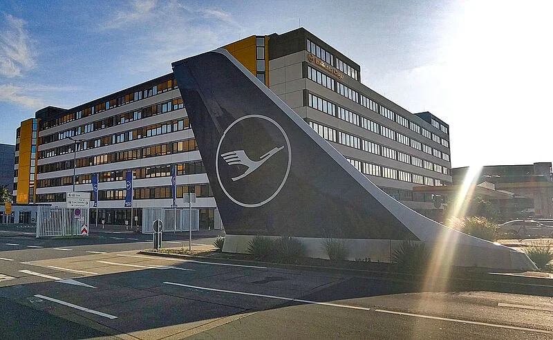 Bürogebäude der Lufthansa am Flughafen Frankfurt im Gegenlicht, davor ein aufgestelltes Seitenleitwerk mit dem Kranich-Zeichen.