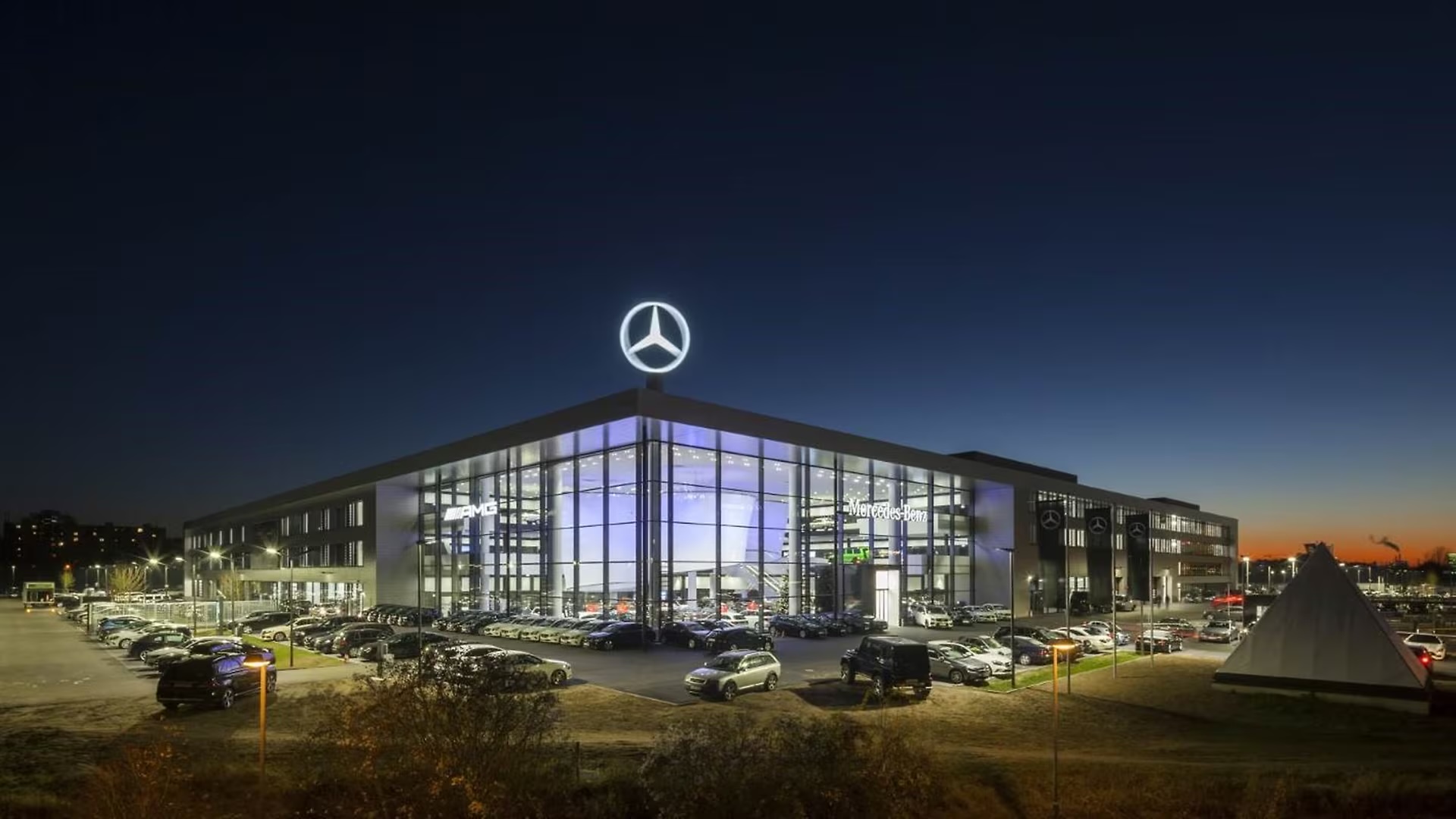 Das Gebäude der Mercedes-Benz Niederlassung Frankfurt am Main in der Abenddämmerung: eine beleuchtete Glasfassade über zwei Geschossen, auf dem Dach der leuchtende Stern, davor ein voll belegter Stellplatz.