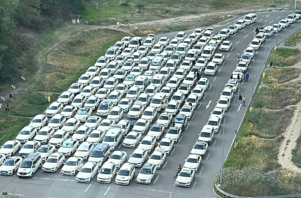 Luftaufnahme des Sammelplatzes am Flughafen: rund 150 weiße Taxis stehen in geordneten Reihen auf einer asphaltierten Fläche, rechts daneben eine Zulaufspur mit einer weiteren Wagenschlange.